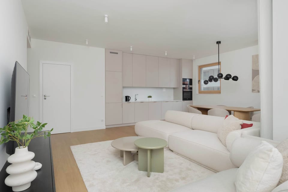 An open-plan living and dining space with a round table and lounge area. Large windows provide abundant daylight throughout the room.