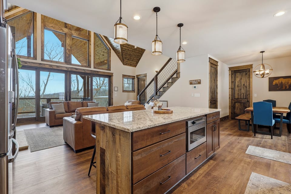 A modern rustic kitchen with wood cabinetry, stainless steel appliances, a central island with seating, pendant lighting, white tile backsplash, and wide plank hardwood flooring.