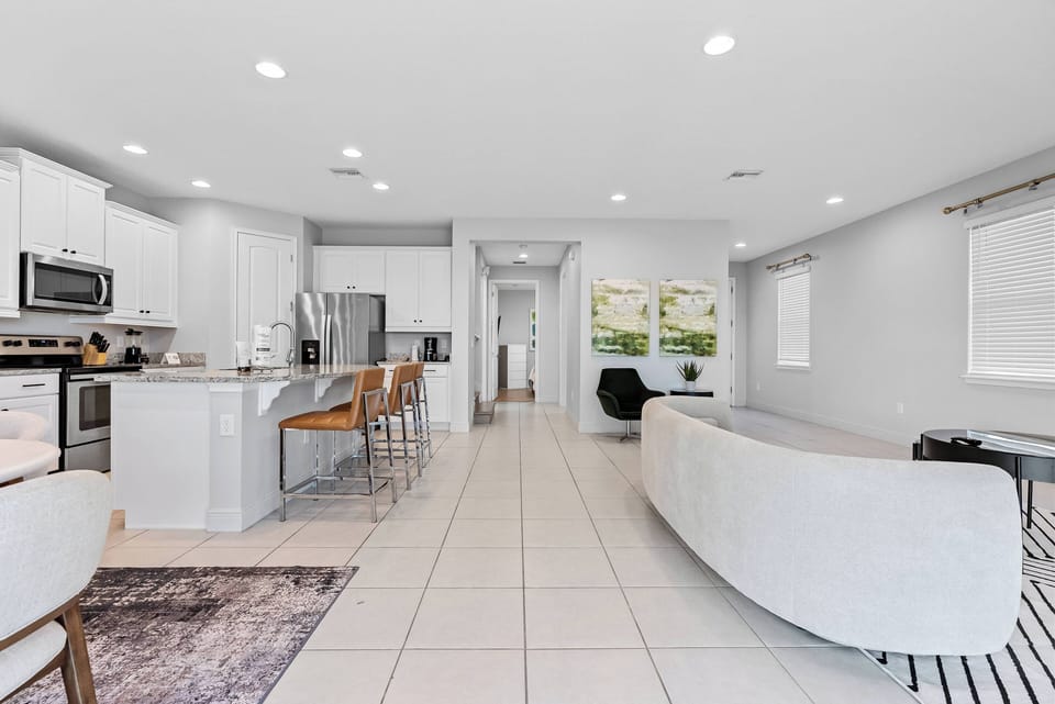Experience the bright, open-concept living area featuring sleek white cabinetry and polished tile flooring that amplify natural light. The cozy curved sofa and chic bar stools create a perfect spot for relaxation and gatherings.