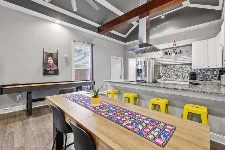 Dining area with a long table, breakfast-bar seating, nearby shuffleboard table, and open kitchen.