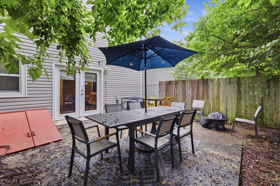 Reverse patio view with a black dining table, umbrella, multiple chairs, rear glass doors, privacy fence, and surrounding trees.