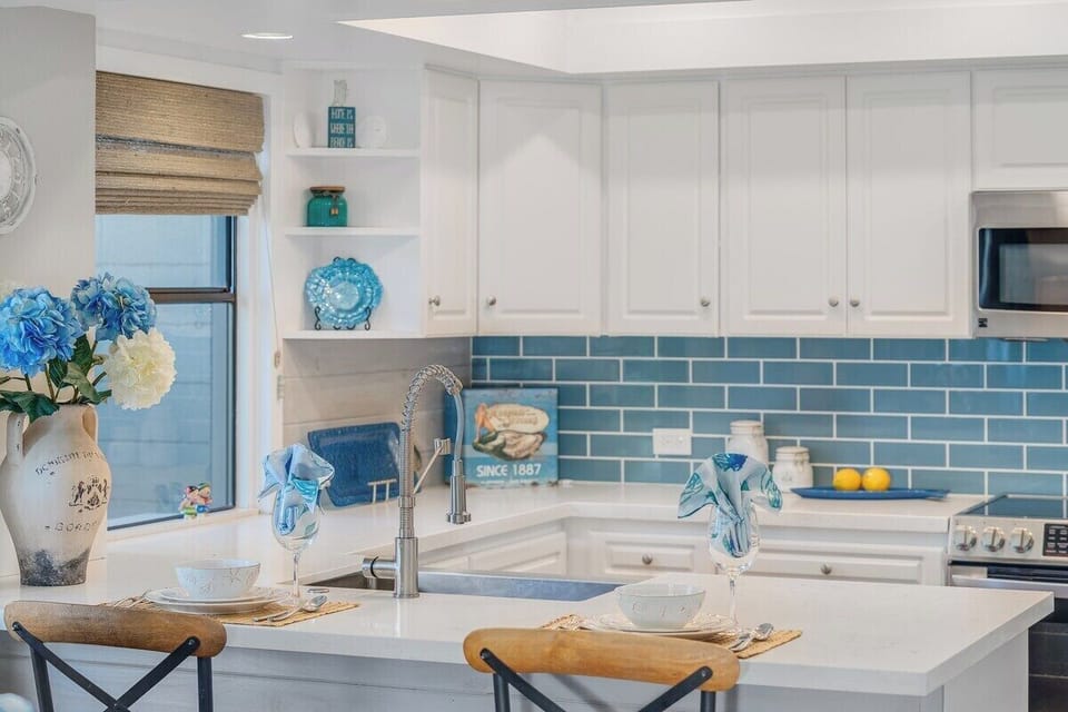 Prepare meals in a bright kitchen featuring white cabinetry, a sleek farmhouse sink, and a stunning blue tile backsplash accented by coastal décor.