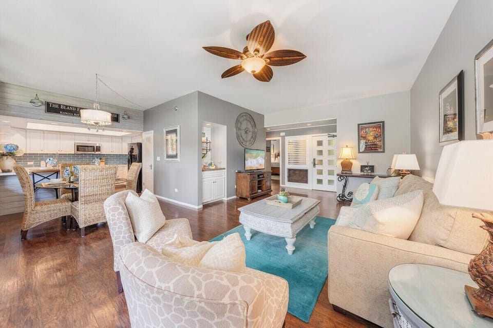 Relax in a spacious open-concept living room featuring neutral-toned furniture, tropical accents, and a ceiling fan for added comfort.