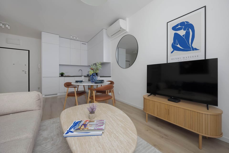 A living and dining area with a beige sofa and a round coffee table. The kitchen area is visible in the background, creating an open-plan layout.