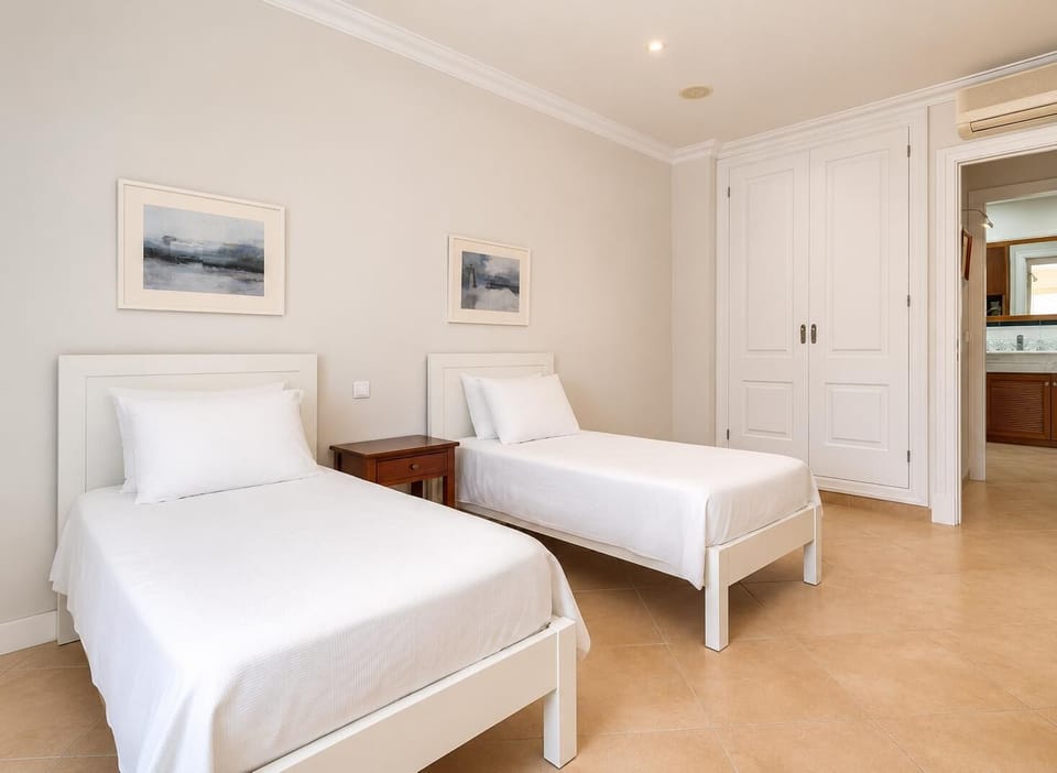A clean and modern guest room featuring two single white beds, a central wooden nightstand, and large built-in wardrobes.