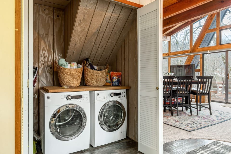A laundry closet contains a front-loading washer and dryer beneath a wood counter with woven supply baskets.