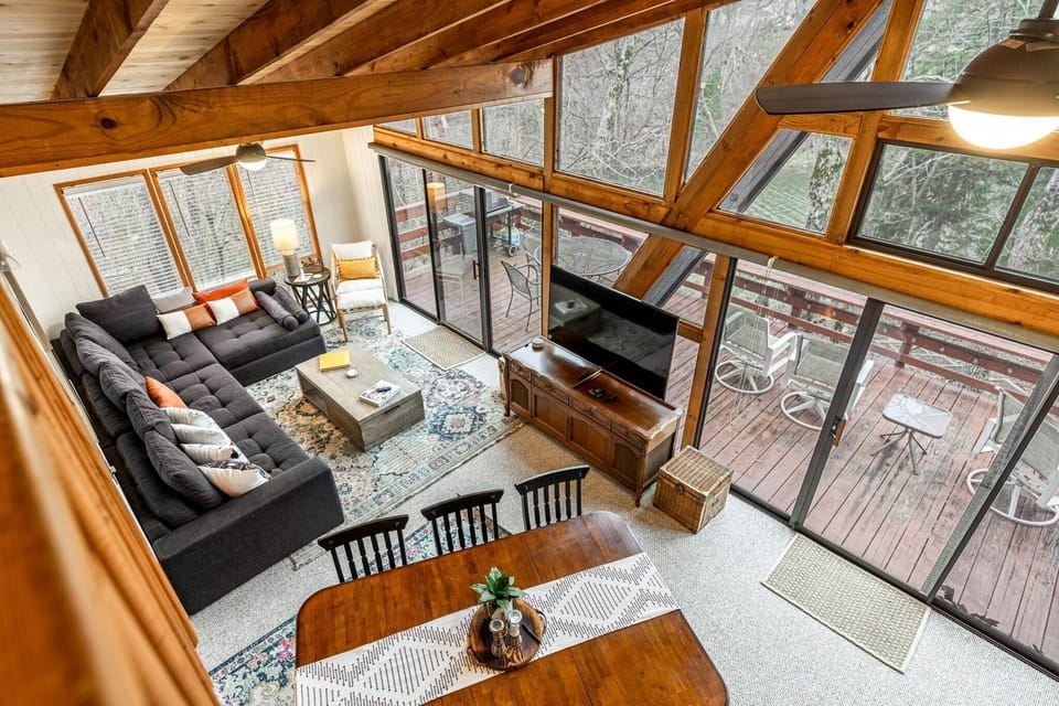 An elevated view shows the open living and dining area beneath exposed beams, with floor-to-ceiling windows and the furnished deck beyond.