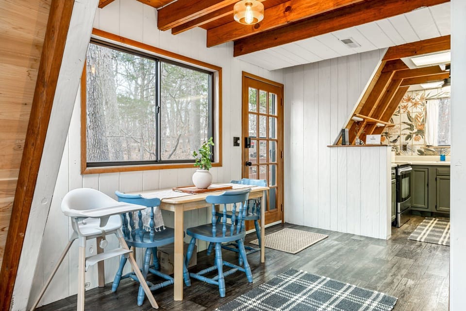 A light wood table with blue chairs and a high chair sits beside the glass deck door, with the kitchen visible through the adjoining opening.