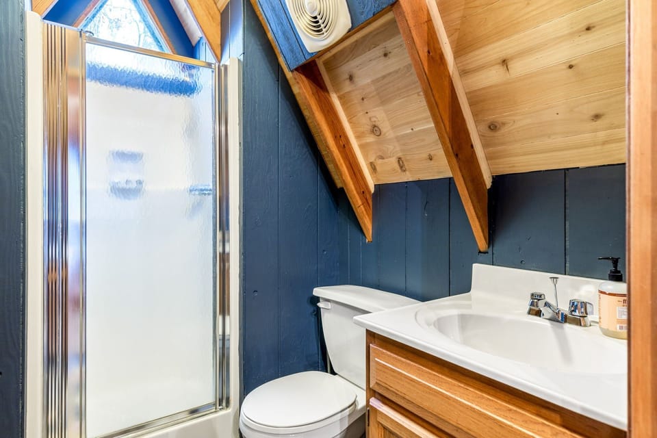 A compact bathroom beneath the angled roof includes a frosted-glass shower, toilet, and wood vanity with a white sink.