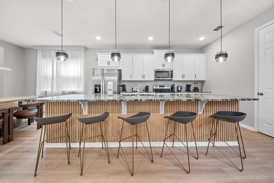 Join us at our chic glass-topped island, where modern bar stools offer a relaxed spot for dining and chatting. The bright kitchen, with white cabinetry and warm wood accents, is beautifully lit by large windows that bring in.