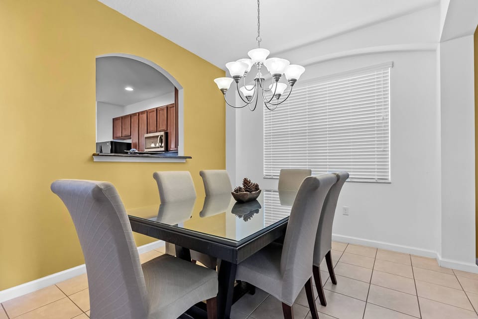Experience meals in this stylish dining area, featuring a sleek glass table surrounded by plush gray chairs. The warm yellow wall adds a cheerful touch, while a modern chandelier casts a soft glow, creating a welcoming atmosphere.