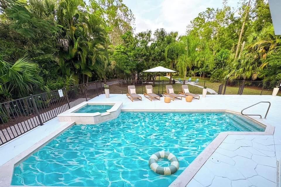 Step into my Naples, Florida oasis: a sunlit pool framed by palms and garden greenery, plush loungers and pool beds inviting calm, with a soothing spa and bubbling jacuzzi warming beside the water.