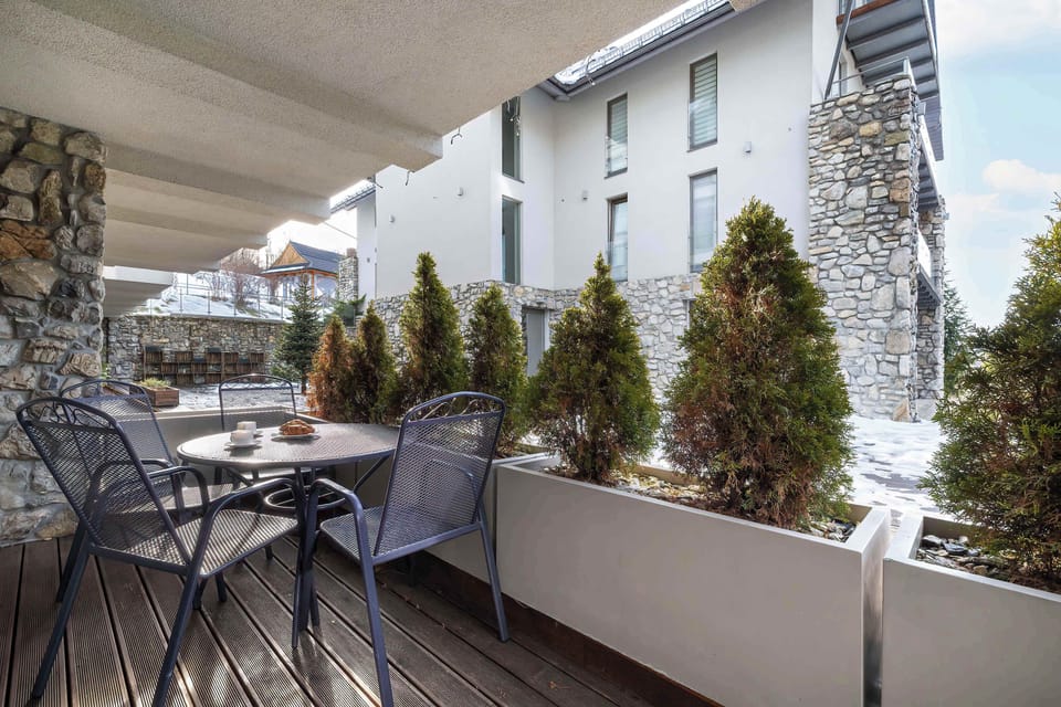 A balcony view showing outdoor seating and surrounding apartment buildings. Decorative plants line the railing. The space feels private and pleasant.