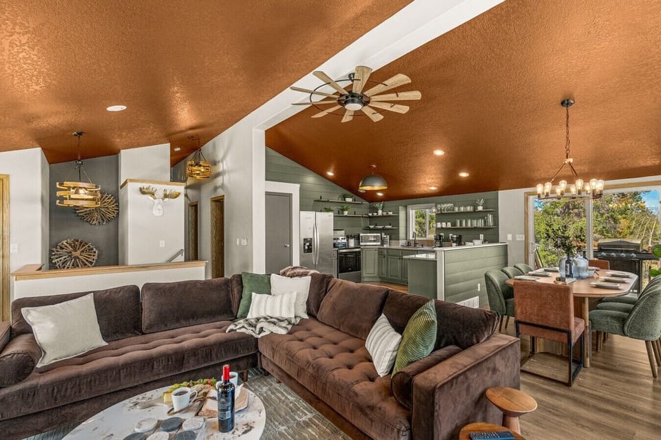 Open-concept mountain chalet great room combining a large sectional sofa, dining area set for six, and fully equipped kitchen. Features vaulted copper-toned ceiling, rustic windmill ceiling fan, and modern decor in Fairplay, CO.