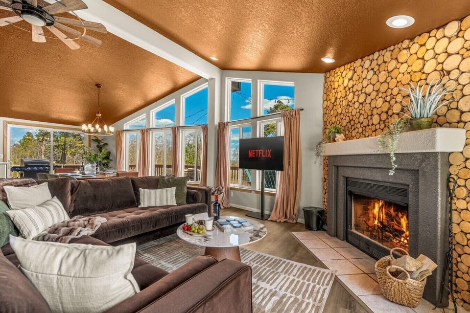 Modern open living room with floor-to-ceiling windows, mountain views, a cozy wood-burning fireplace, and spacious seating for groups. Perfect for relaxing after exploring Breckenridge, Fairplay, and nearby hiking, skiing, and outdoor adventures.