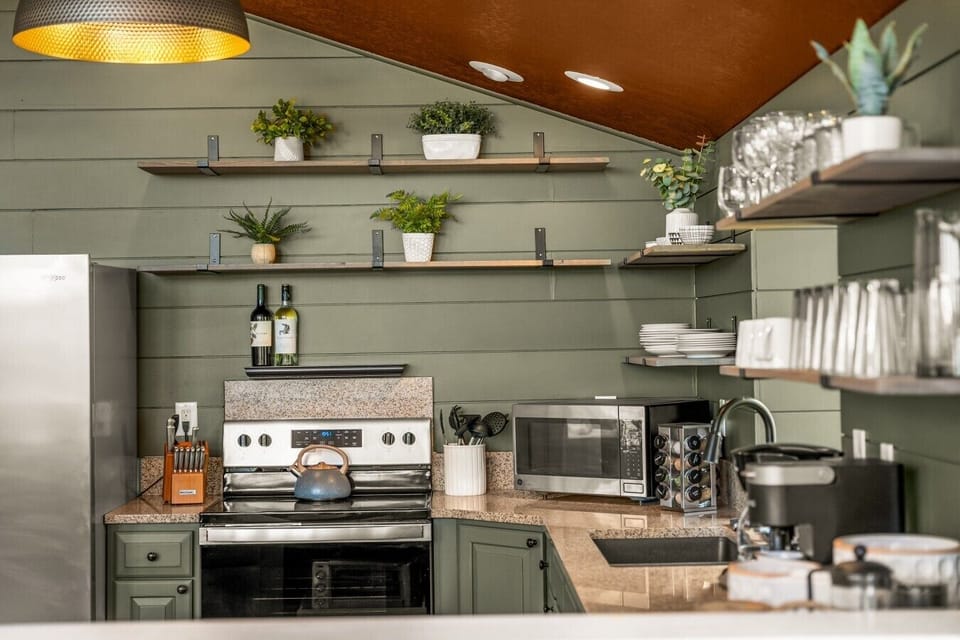 Modern cabin kitchen corner featuring green shiplap walls, open wooden shelves with glassware, stainless steel stove, microwave, coffee maker, and granite countertops. Fully stocked chef kitchen in Fairplay, CO cabin near Breckenridge.