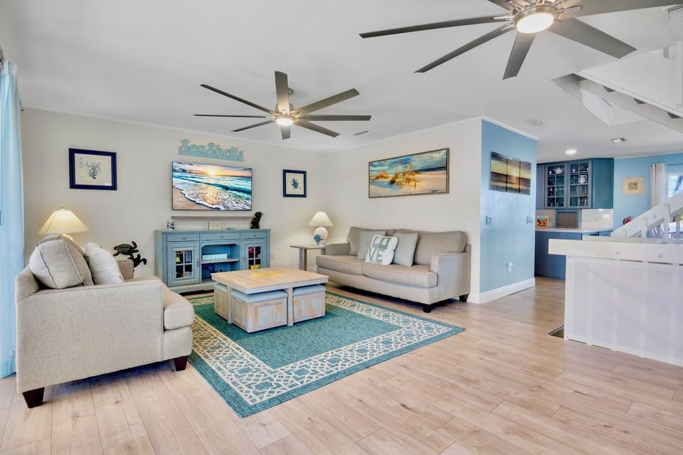 Sink into plush sofas beneath gentle ceiling fans while coastal artwork and turquoise accents create your perfect beach retreat atmosphere.