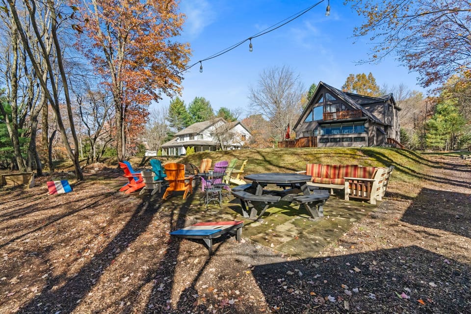 Spacious backyard with colorful Adirondack chairs, a picnic table, cushioned bench, cornhole, and overhead string lights - a fun gathering spot set against the wooded hillside and cabin