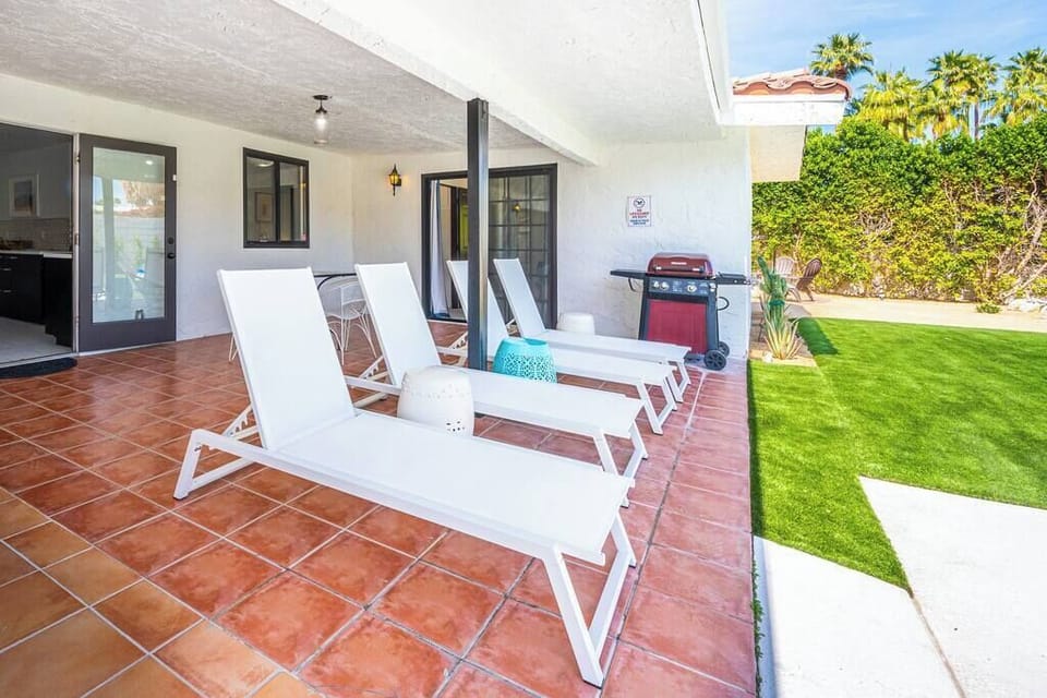Patio lounge area with four white sun loungers, gas grill, and lush turf lawn — your afternoon lineup sorted