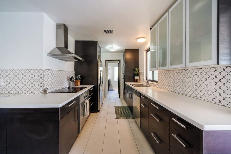 Galley kitchen with quartz counters, arabesqe tile backsplash, stainless hood, and glass-front upper cabinets — well-equipped for real cooking