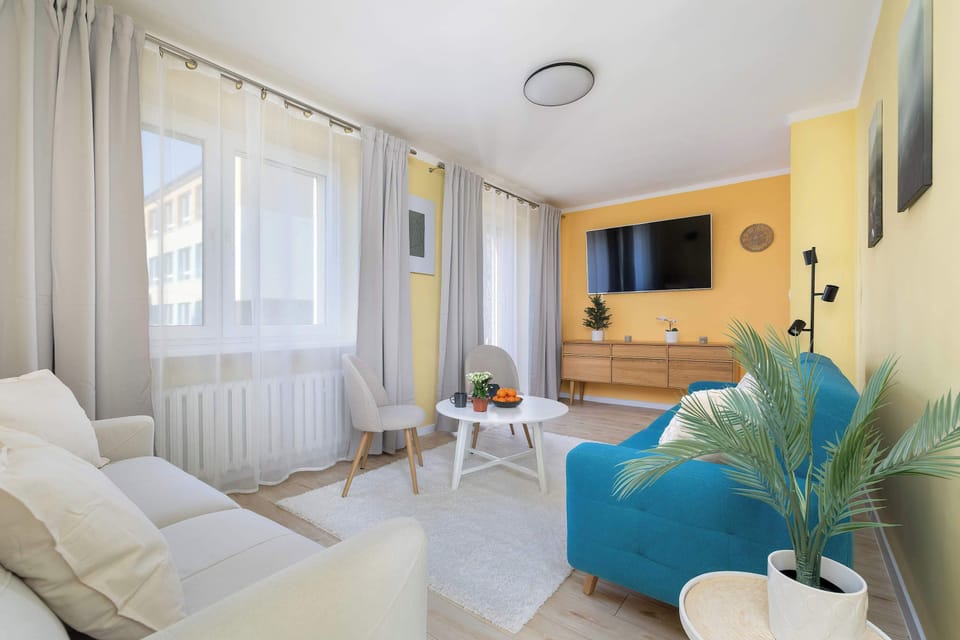 A cozy living area with soft seating and sheer curtains covering large windows. Plants bring a natural touch to the space. The room feels warm and inviting.