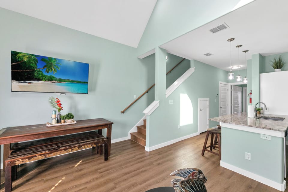 Your open-plan living space features soothing sage walls, tropical artwork, and a kitchen island perfect for morning coffee.