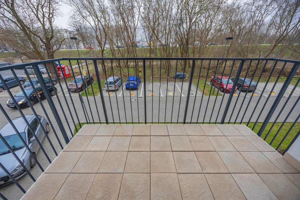 A balcony with metal railings overlooking a green outdoor area. The floor is tiled and well-kept. The space offers fresh air and an open view.
