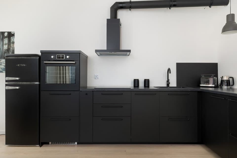 A sleek black kitchen with matte cabinets, a built-in oven, and exposed ventilation piping. The design feels industrial and modern. Clean surfaces dominate the space.