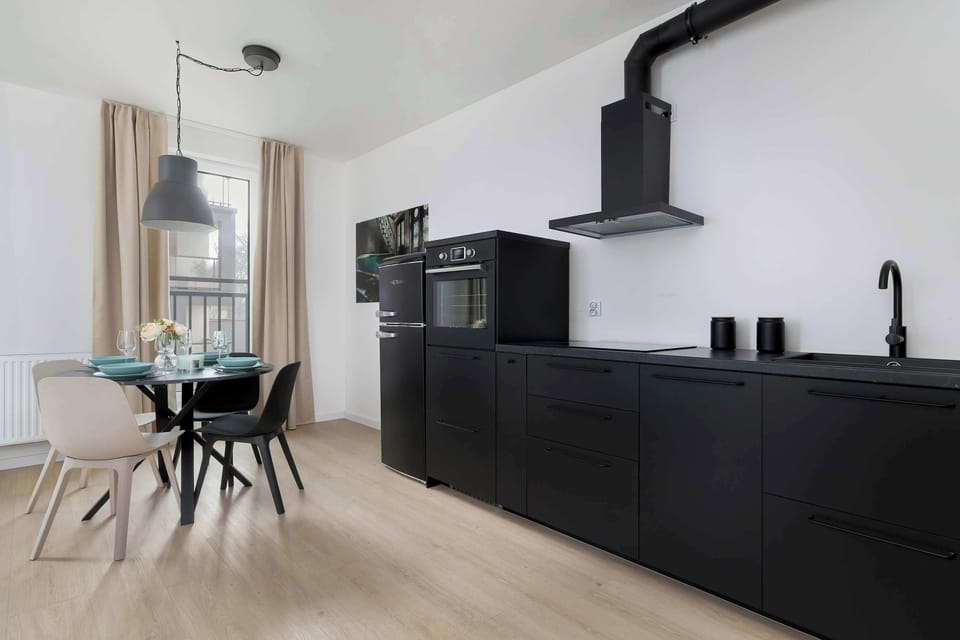 A black kitchen with a dining table placed close to the cooking area. Modern lighting fixtures hang from the ceiling. The space blends functionality with style.