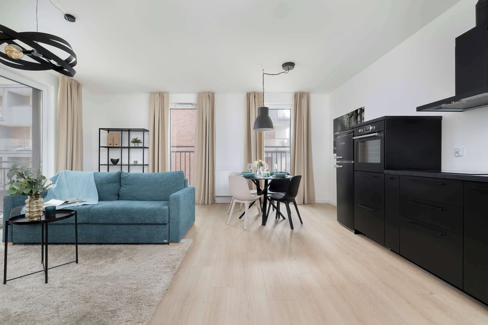 A wide view of the open-plan living room and kitchen with light wood flooring throughout. The blue sofa contrasts with the black kitchen cabinets. The space feels airy and modern.