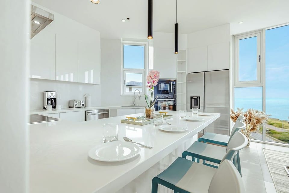 A bright, modern kitchen opens to stunning coastal vistas through floor-to-ceiling windows. Sleek white surfaces and contemporary fixtures create a refined space for preparing meals while enjoying breathtaking ocean views.