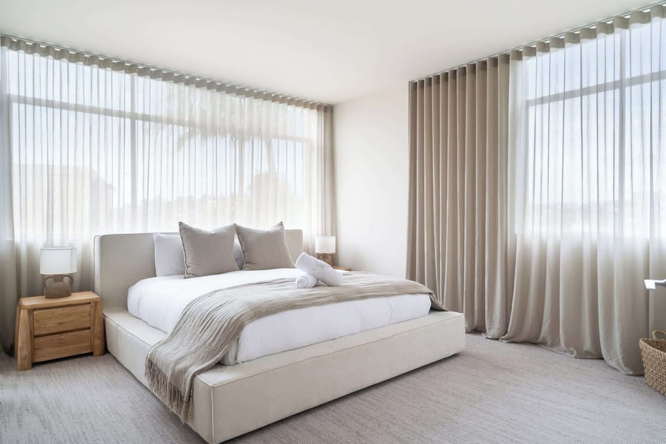 Elegantly appointed master retreat featuring a luxurious king bed with plush bedding, expansive built-in wardrobes, and sheer curtains framing a tranquil leafy outlook. including room-darkening shades ensure a restful nights sleep