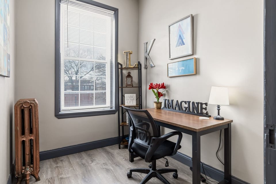 A dedicated workspace offers a desk, office chair, lamp, artwork, radiator, and abundant natural light.