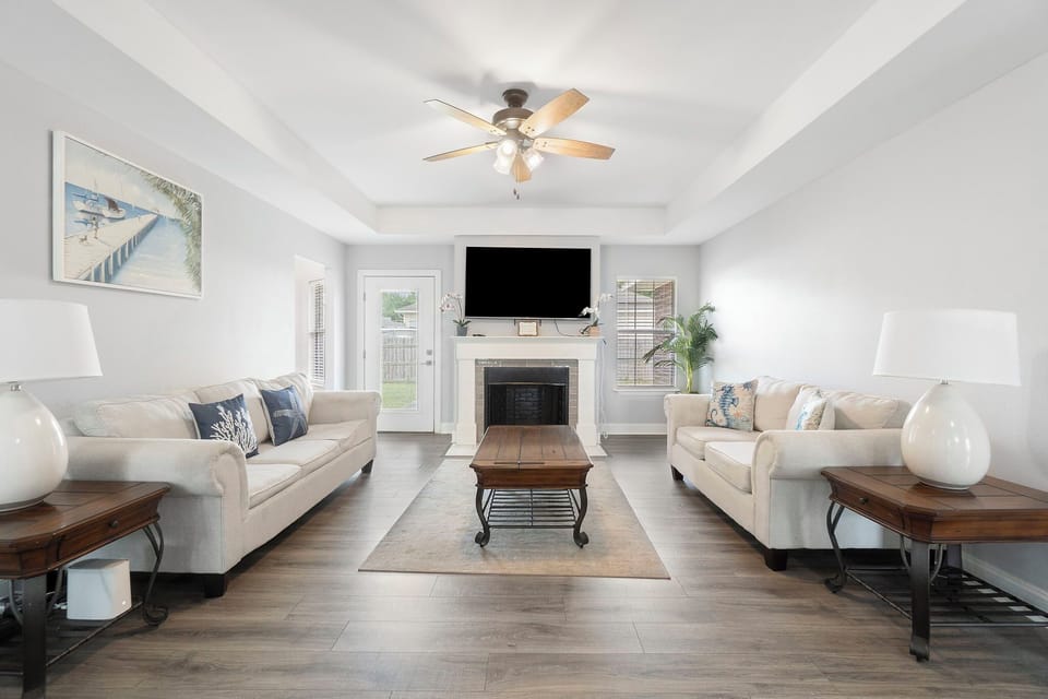 Step into comfort in this spacious living room, featuring matching sofas, a stylish ceiling fan, and a cozy fireplace as the centerpiece. Natural light pours in from multiple windows and a glass door, creating the perfect setting for relaxation.