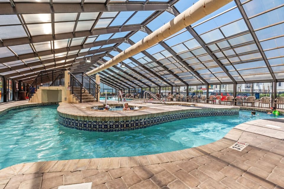 Indoor pool area enclosed by a bright glass structure with windows all around and overhead. Features a year-round pool, lazy river, and hot tub, creating an airy, light-filled resort atmosphere.