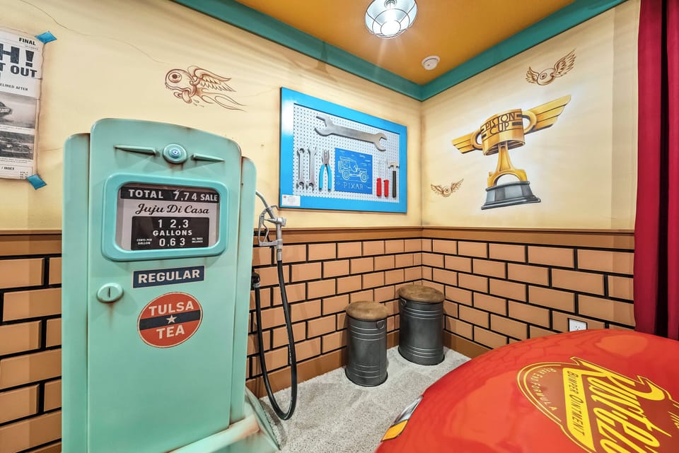 Enter a vibrant room filled with a playful retro vibe, featuring a turquoise gas pump and nostalgic artwork. The warm orange ceiling and charming brick accents create an inviting atmosphere ideal for relaxation and fun.