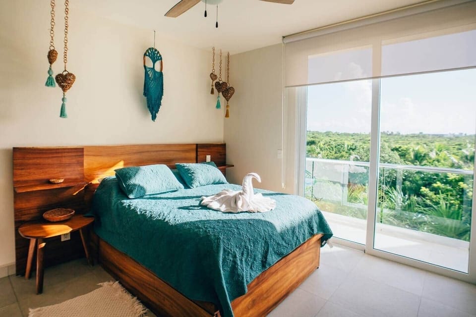 The second bedroom features a comfortable queen-size bed, a private balcony with Akumal jungle views, air conditioning, a ceiling fan, room-darkening shades, power sockets by the bed, and easy-access light switches.