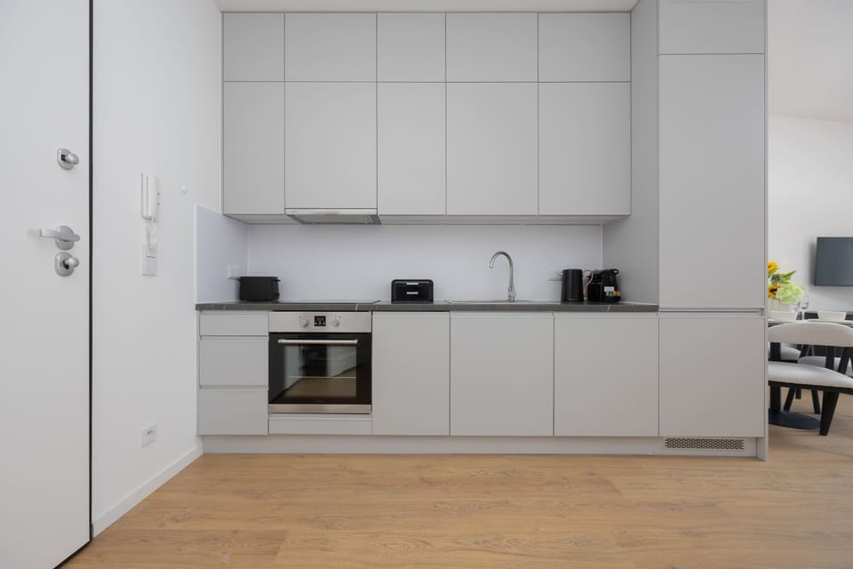 A wide kitchen view showing white cabinets, built-in oven, and light flooring. The layout appears efficient and modern. Everything looks well-organized.