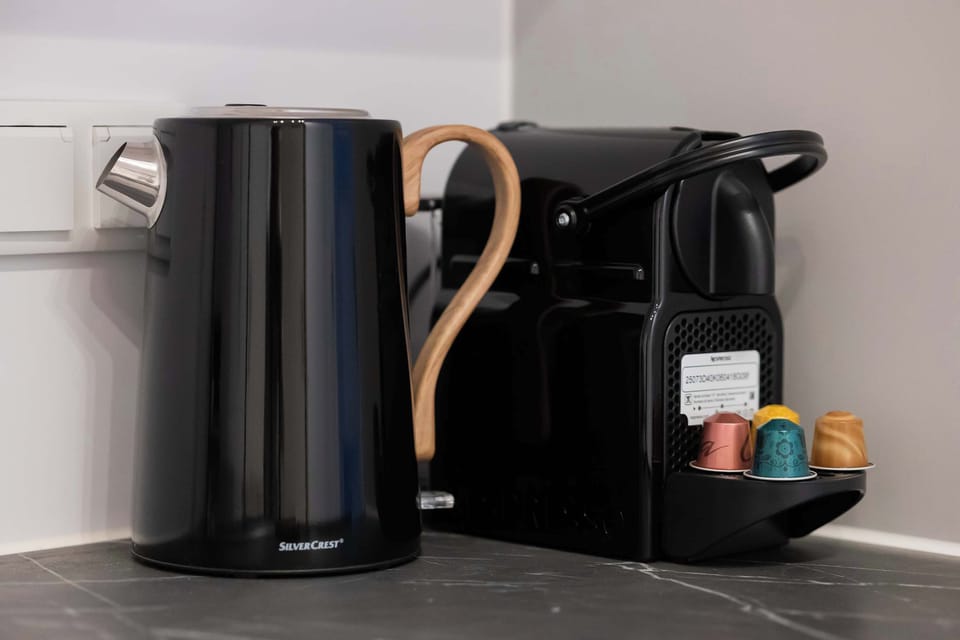 A close-up of a kettle and coffee setup placed on a dark countertop. Cups and accessories are neatly arranged. The space looks ready for daily use.