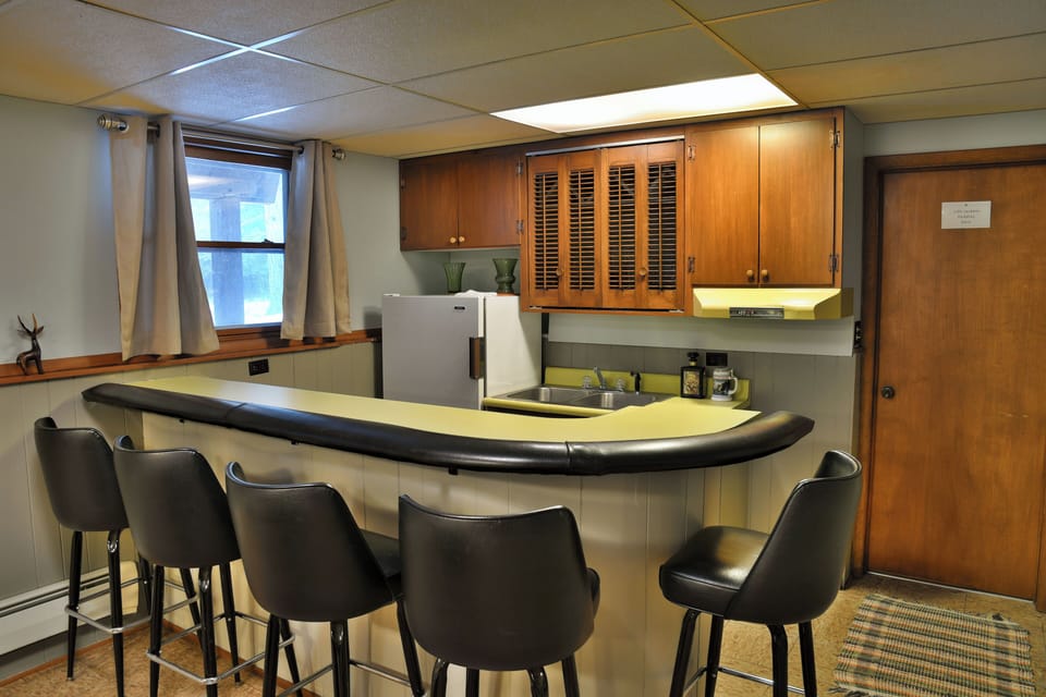 Lower-level bar at Elk Crossing Lakehouse with a curved yellow countertop, six black barstools, wood cabinets, and a small kitchenette area.