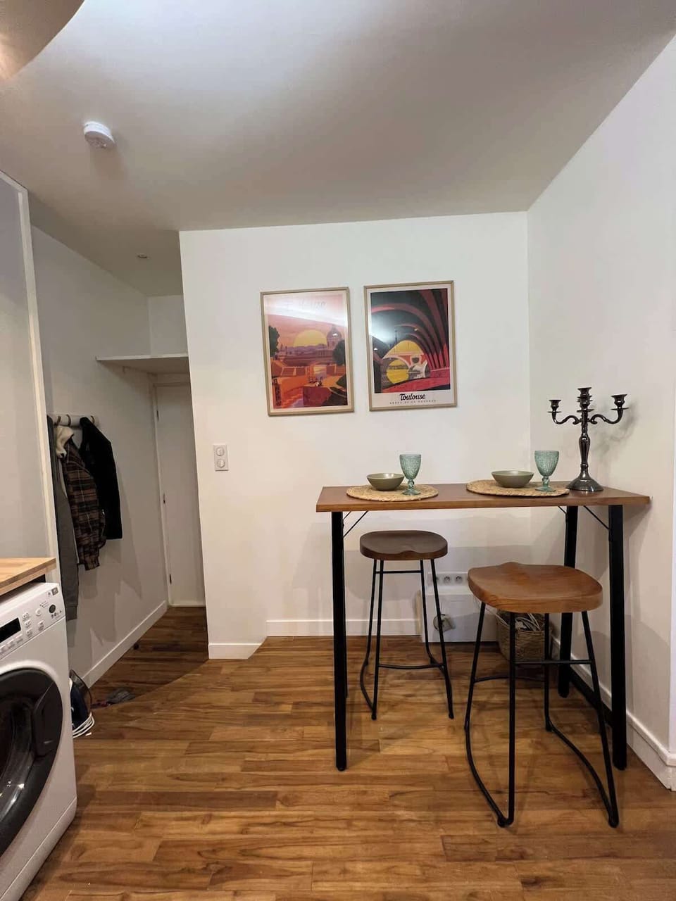 Coin cuisine avec table haute, tabourets et tableaux — lave-linge visible et parquet au T2 Le Câlin