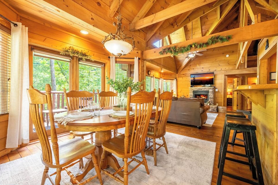 Gather around the handcrafted wood table in this warm, cathedral-ceiling space where sunlight streams through forest views and a stone fireplace awaits your evenings together.