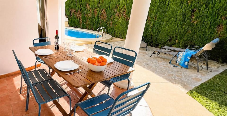 This sun-drenched Mediterranean villa terrace features an outdoor wooden dining table set for al fresco meals next to a private swimming pool and a sun-soaked lounging patio.