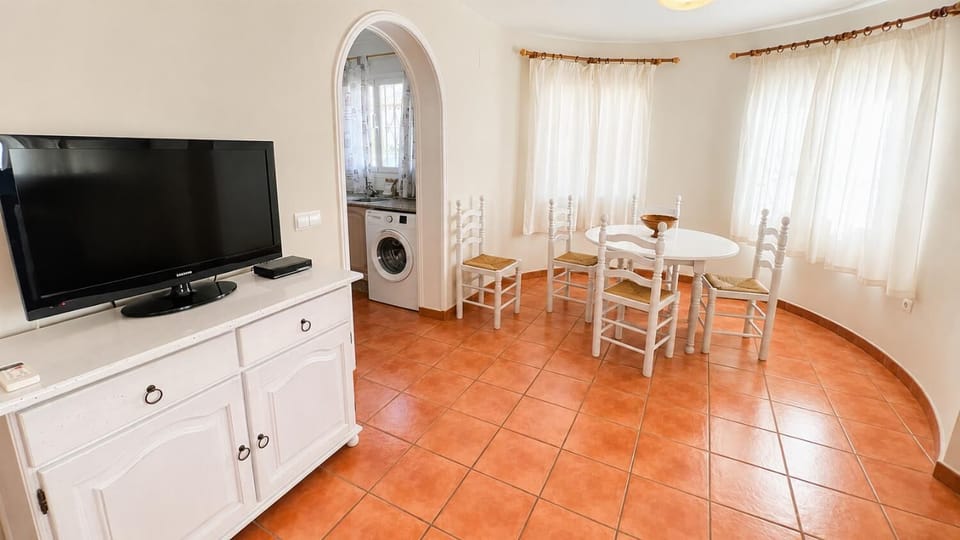 This charming Mediterranean holiday villa features a sunlit open-plan dining area with rustic terracotta flooring, a flat-screen TV setup, and convenient adjoining kitchen access for a seamless self-catering stay.