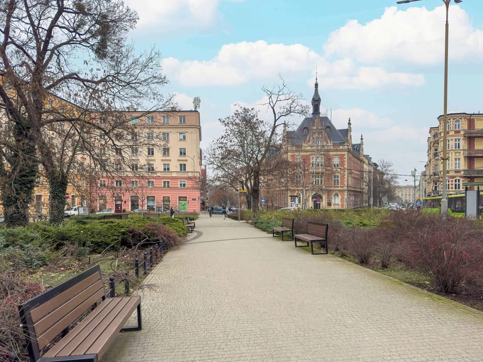 Green outdoor walkway with park benches, leafless trees, and historic city buildings.