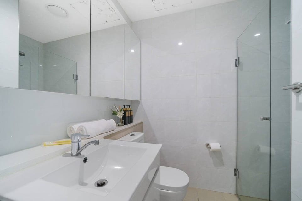 Second bathroom with a walk in shower behind a glass door, a toilet and a wide vanity with a mirrored cabinet. Towels and toiletries sit on the ledge.