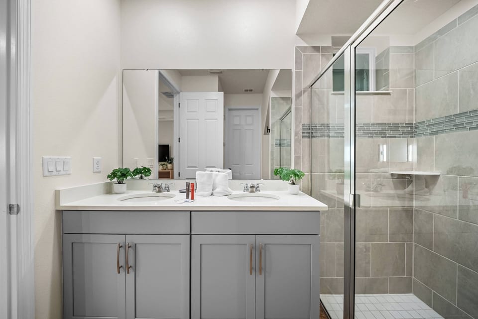 Experience a beautifully designed bathroom with sleek gray cabinetry and a spacious double vanity. Enjoy the elegant tile work in the glass shower and the natural light that creates a bright, refreshing atmosphere for your stay.