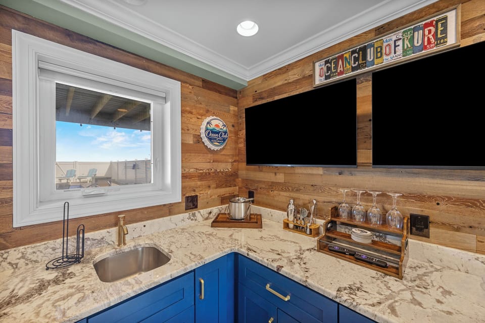 Your coastal-inspired bar awaits with dual flatscreens, elegant glassware, and granite counters—perfect for mixing drinks while gazing at ocean views through the window.