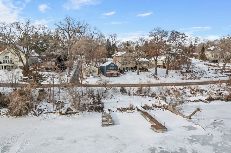 A peaceful lakefront neighborhood blanketed in pristine snow, featuring charming homes nestled among bare winter trees beneath a bright blue sky.