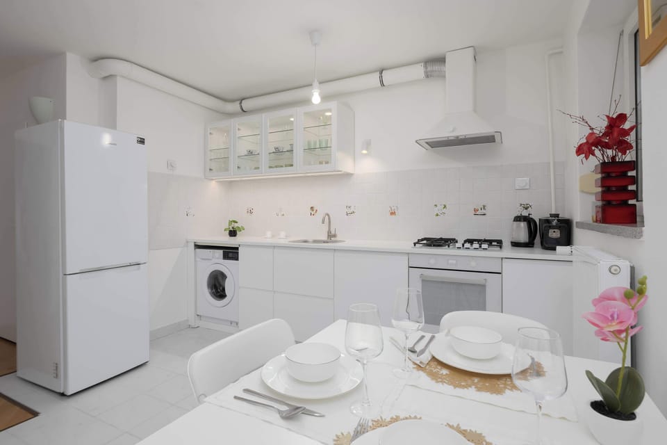 Kitchen interior featuring white cabinets, a fridge, oven, and neatly arranged tableware. The dining area is visible in the corner.